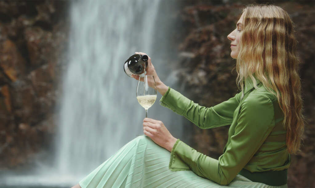 Wide shot of a woman pouring a Bonterra white wine into a wine glass. Waterfall in the background.